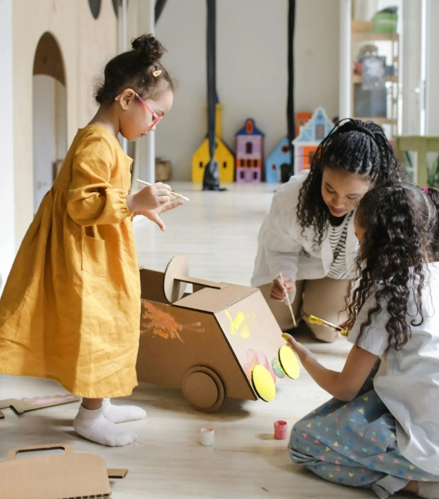Two children and caregiver crafting and playing with cardboard car in bright playroom, inspiring creativity.
