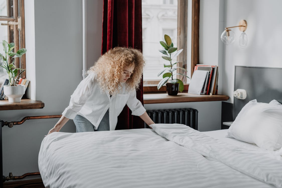 Person making bed in bright tidy bedroom, symbolizing order, comfort, and nurturing environment.